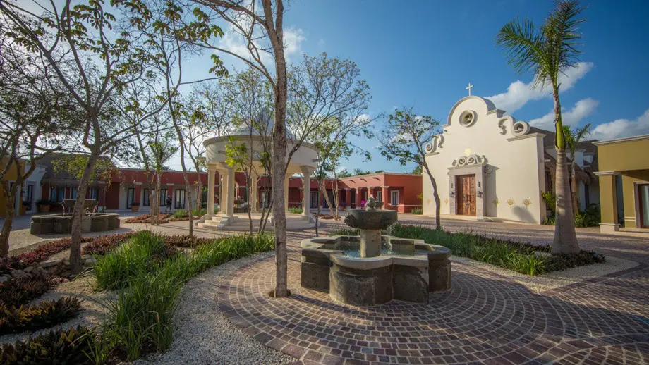 The El Pueblito chapel plaza at Mayakoba: a white chapel with a cross on its scrolled gable and carved wooden doors, beside a domed rotunda and a stone fountain on brick paving, framed by palms