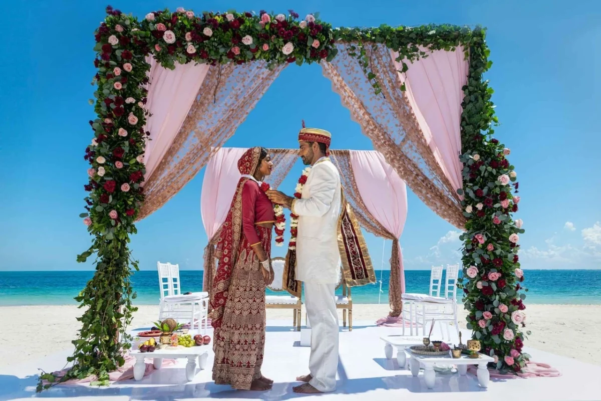 Couple in red lehenga and white sherwani under a floral mandap on the beach at Grand Palladium Costa Mujeres