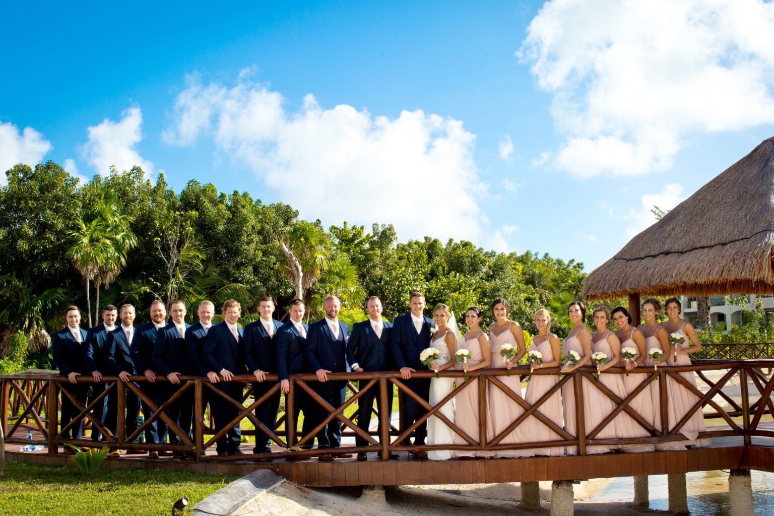 A bride and groom in a navy suit embrace on a wooden bridge framed by palms at The Grand at Moon Palace Cancun