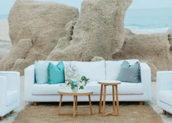 White lounge seating with turquoise and gray pillows on the beach at Grand Velas Los Cabos, set against rock formations and the ocean