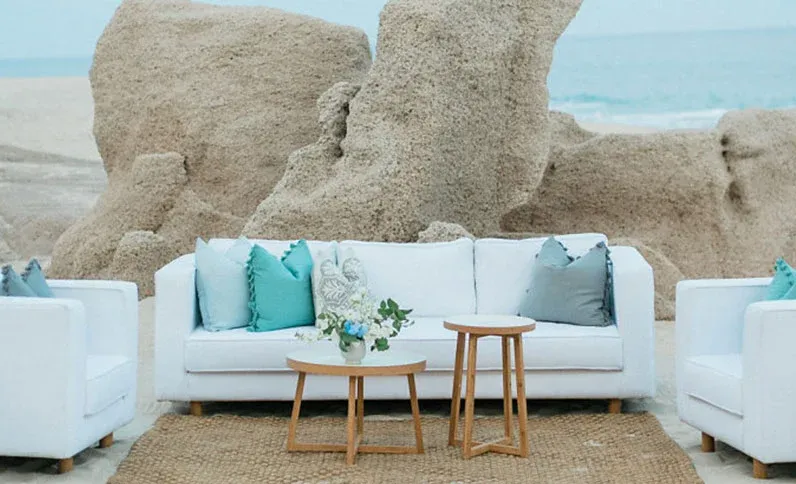 White lounge seating with turquoise and gray pillows on the beach at Grand Velas Los Cabos, set against rock formations and the ocean