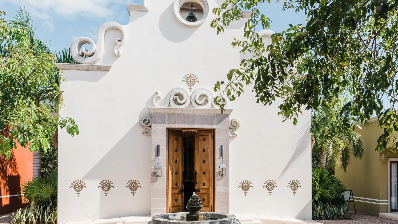 The white chapel facade at The Little Town, Rosewood Mayakoba: a scrolled baroque gable with a bell hanging in a circular opening above carved wooden double doors in a stone surround, with a stone fountain in front
