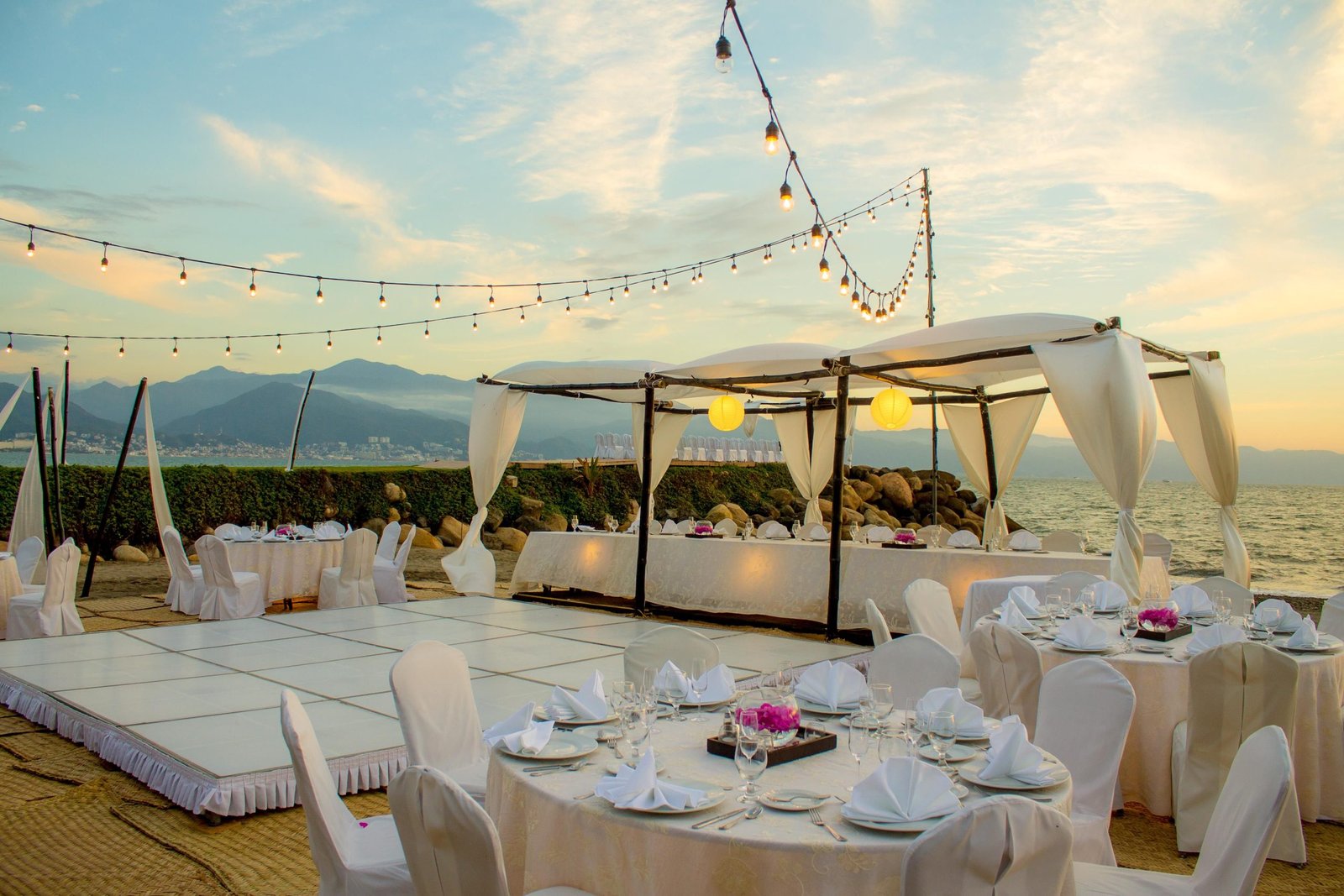 Beachfront wedding reception at sunset at Velas Vallarta with round tables, a white dance floor and festoon lighting over the water